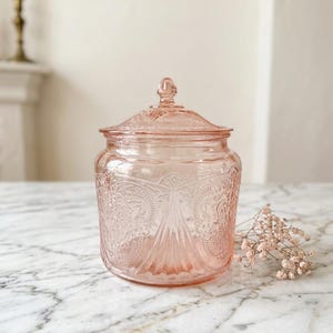 Könnte beinhalten: Ein rosa Glasbehälter mit Deckel, mit einem geprägten Blumendesign. Der Behälter ist rund mit einem dekorativen Knauf auf dem Deckel. Der Behälter steht auf einer Marmoroberfläche.