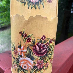 May include: A vintage, yellow metal container with a scalloped top edge. The container is adorned with hand-painted floral designs in shades of pink, red, orange, and green. The container has a distressed, crackled finish.