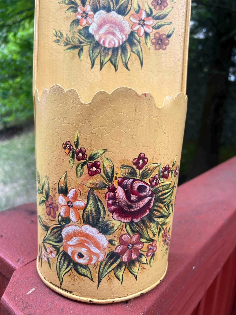 May include: A vintage, yellow metal container with a scalloped top edge. The container is adorned with hand-painted floral designs in shades of pink, red, orange, and green. The container has a distressed, crackled finish.