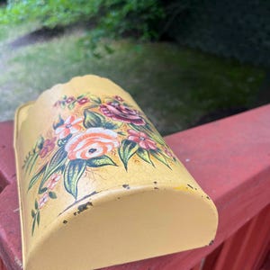 May include: A yellow metal wall pocket with a floral design. The flowers are in shades of pink, orange, and red, with green leaves. The pocket has a curved shape and is slightly distressed.
