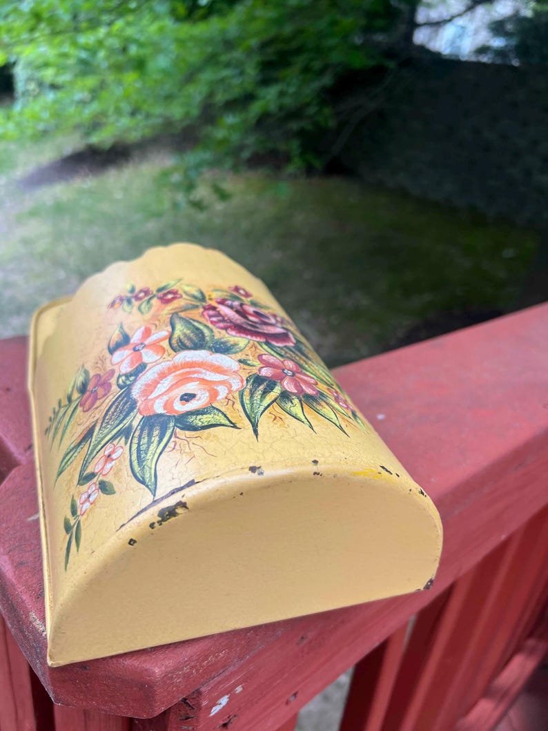 May include: A yellow metal wall pocket with a floral design. The flowers are in shades of pink, orange, and red, with green leaves. The pocket has a curved shape and is slightly distressed.