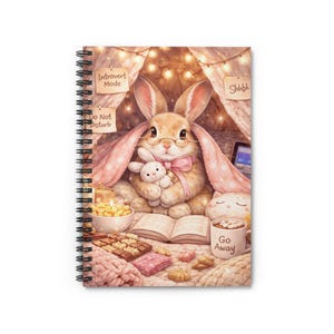 May include: Spiral-bound notebook featuring a whimsical illustration of a bunny in a cozy setting. The bunny wears glasses and holds a stuffed animal. The scene includes a book, snacks, and the text "Introvert Mode" and "Do Not Disturb".