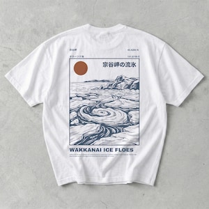 May include: White t-shirt featuring a graphic of the Wakkanai Ice Floes. The design includes a brown sun, a blue and white landscape with text in Japanese and English. The shirt has short sleeves and a crew neck.