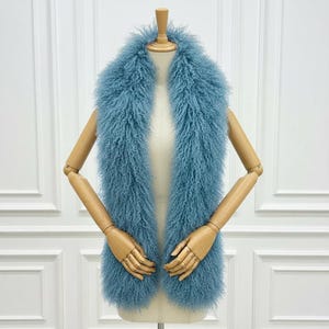 May include: A light blue, fluffy scarf on a mannequin. The scarf is long and rectangular, made of a soft, textured material. The colour is a muted, dusty blue, and the scarf appears warm and cosy, perfect for colder weather.