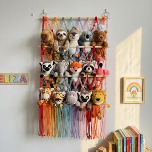 May include: A multi-colored macrame wall hanging organizer featuring a grid of knotted cords and horizontal wooden dowels. The organizer holds a collection of various plush animal toys, including a bear, koala, penguin, elephant, and giraffe.