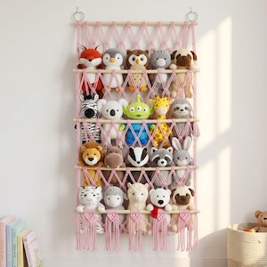 May include: A wall-mounted macrame storage organiser featuring pink knotted cord netting and horizontal wooden dowels. The organiser holds twenty assorted plush animal toys, including a fox, penguin, owl, cat, pug, zebra, koala, alien, giraffe, sloth, lion, hippo, badger, raccoon, rabbit, tiger, sheep, llama, bear, and dog.