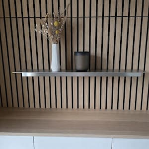 以下が含まれることがあります： 木製のスラット壁と黒いアクセントが特徴のモダンなインテリアデザイン。ステンレス製の棚には、ドライフラワーが入った白い花瓶と濃い灰色のキャンドルが置かれています。下には、白いキャビネットがクリーンなコントラストを加えています。