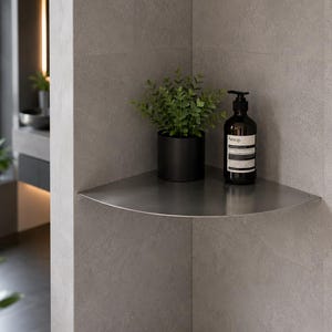 May include: A brushed metal corner shelf mounted on a grey tiled wall. On the shelf sits a small green plant in a matte black cylindrical pot and a dark brown Aesop pump bottle with a white label containing black text.