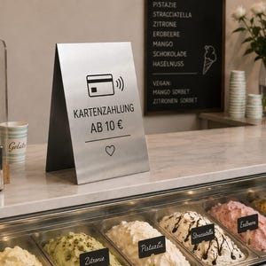 Op de afbeelding: Een roestvrijstalen bord met de tekst "KARTENZAHLUNG AB 10 €" en een creditcardsymbool, dat betalingsopties aangeeft. Het bord staat op een marmeren toonbank in een ijssalon.