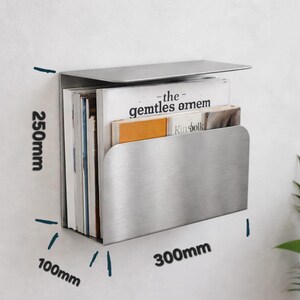 May include: A brushed stainless steel wall-mounted magazine rack. The rack is 300mm wide, 100mm deep, and 250mm high, holding several magazines. The top of the rack folds over. The magazines include the text "the gentles amem" and "Kinfolk".