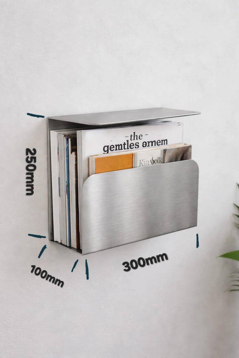 May include: A brushed stainless steel wall-mounted magazine rack. The rack is 300mm wide, 100mm deep, and 250mm high, holding several magazines. The top of the rack folds over. The magazines include the text "the gentles amem" and "Kinfolk".
