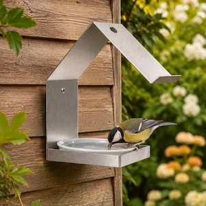 May include: A wall-mounted bird feeder made of brushed stainless steel with a peaked roof design and a circular white ceramic dish for water or seeds. A small yellow, black, and white bird is perched on the edge of the dish drinking water.