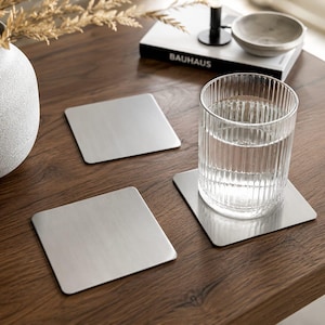 Könnte beinhalten: Drei quadratische Untersetzer aus gebürstetem Metall auf einem Holztisch. Ein klares Wasserglas steht auf einem Untersetzer. Eine dekorative Vase und ein Buch mit dem Titel "BAUHAUS" befinden sich ebenfalls auf dem Tisch. Die Untersetzer sind etwa 10 cm groß.