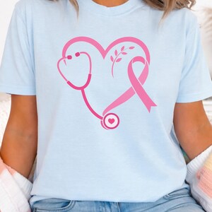 May include: A light blue short-sleeve t-shirt featuring a pink graphic on the chest. The design consists of a stethoscope and a breast cancer awareness ribbon arranged to form a heart shape, with a small sprig of leaves inside the heart outline.