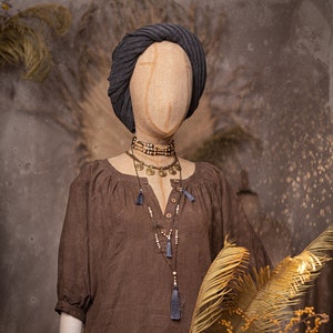 Mannequin wearing one brown linen boho dress with gathered neckline, short sleeves, and loose maxi-length fit; styled with layered necklaces and gray headscarf. Displayed upright, holding dried bouquet. Linen summer dress with pockets.