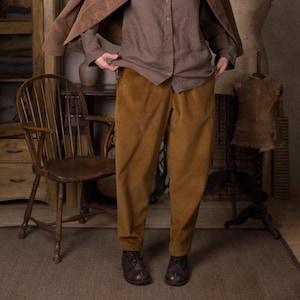 Pode incluir: Um blazer de veludo cotelê castanho, uma camisa de botões castanha com gola redonda e calças de veludo cotelê amarelo-mostarda. O conjunto inclui um chapéu fedora de feltro castanho e sapatos de couro castanho escuro, em tons terra.