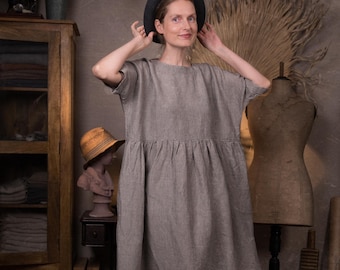 May include: A woman wearing a gray linen dress with a black hat. The dress has a gathered waist and short sleeves. She is standing in front of a wooden cabinet and a mannequin.