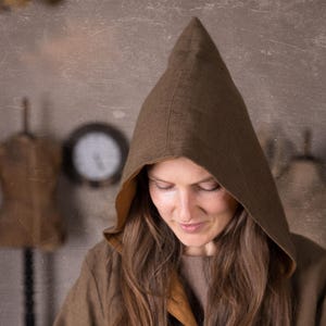 Könnte beinhalten: Ein brauner Kapuzenmantel mit spitzer Kapuze und einem kontrastierenden beigefarbenen Futter. Der Umhang hat einen Knopfverschluss und besteht aus einem strukturierten Stoff. Die Person hat lange braune Haare.