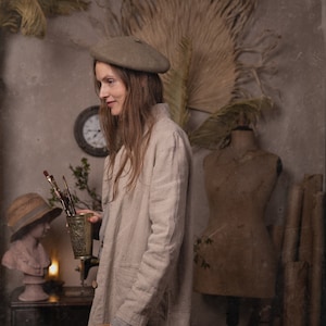 May include: A woman in a light beige linen coat and beret, holding a brass cup of paintbrushes and a wooden picture frame. The studio setting includes a bust, dress form, and a clock, creating a vintage aesthetic.