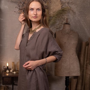 Dark taupe linen dress worn on a model, front three-quarter crop, with round gathered neckline, short button placket, elbow-length puff sleeves, loose body, and visible center front seam.