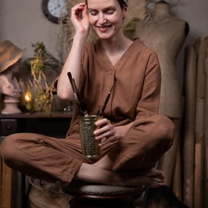 A smiling girl is sitting on a round vintage chair, holding a brass glass with paintbrushes. She is also wearing the 'Artist' jumpsuit from Knock Knock Linen