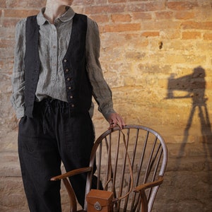 Linen pants in dark charcoal, shown on a model in a three-quarter front view, with a gathered elastic waistband, visible drawstring ties, loose legs, softly wrinkled fabric, and the model standing beside a wooden chair, Knock Knock Linen.