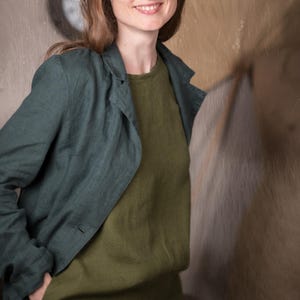 Op de afbeelding: Een vrouw draagt een donkergroene blazer over een olijfgroene top, gecombineerd met een zwarte hoed. De outfit is eenvoudig en stijlvol, met de nadruk op natuurlijke stoffen en een relaxte pasvorm. De achtergrond is neutraal.