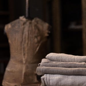 May include: A stack of five folded pieces of fabric in varying shades of beige, taupe, and light gray. The textiles feature a visible, coarse woven texture with frayed edges, stacked neatly in a pile with a blurred dress form in the background.