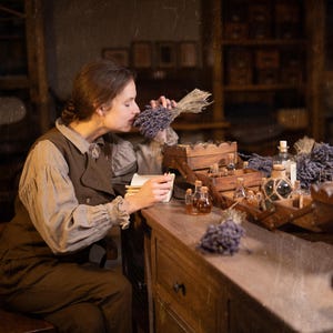 SPARROW linen vest in brown, displayed on a seated model in a side-profile mid-length view, with a sleeveless shape, curved front opening, layered over a light gray collared blouse and brown trousers, beside glass bottles and wooden trays, Knock Knock Linen.