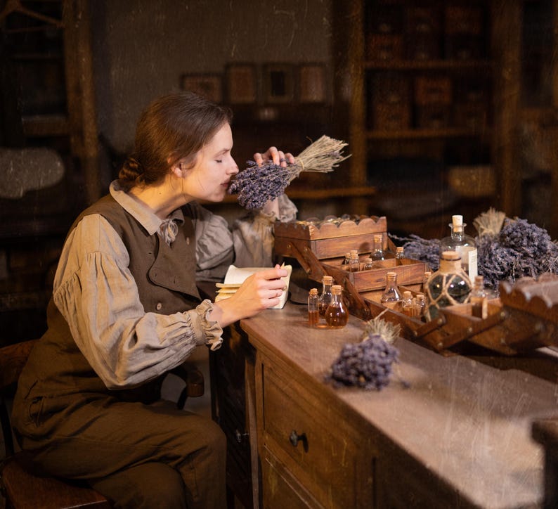 SPARROW linen vest in brown, displayed on a seated model in a side-profile mid-length view, with a sleeveless shape, curved front opening, layered over a light gray collared blouse and brown trousers, beside glass bottles and wooden trays, Knock Knock Linen.