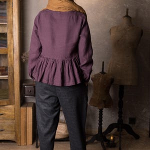 Back view of a woman indoors wearing a plum long sleeve linen peplum blouse with a gathered hem, styled with grey trousers, brown scarf and black hat.