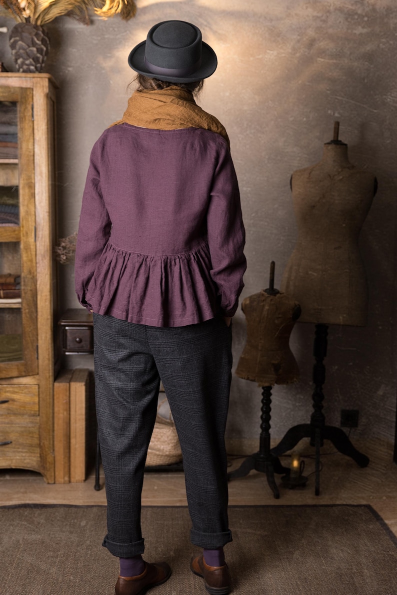 Back view of a woman indoors wearing a plum long sleeve linen peplum blouse with a gathered hem, styled with grey trousers, brown scarf and black hat.