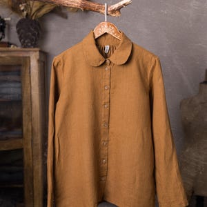 May include: A long-sleeved shirt in a solid mustard-brown color featuring a rounded Peter Pan collar and a front button-down closure with seven matching buttons. The garment is displayed on a wooden hanger, showcasing a simple, straight-cut design.