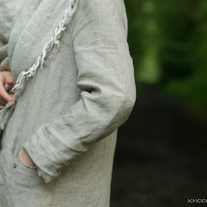 Close-up of a natural linen buttoned dress coat with long sleeves, worn by a person standing outdoors. One hand is tucked into a pocket; the outfit includes a matching linen scarf and a bracelet. Background features soft-focus greenery.