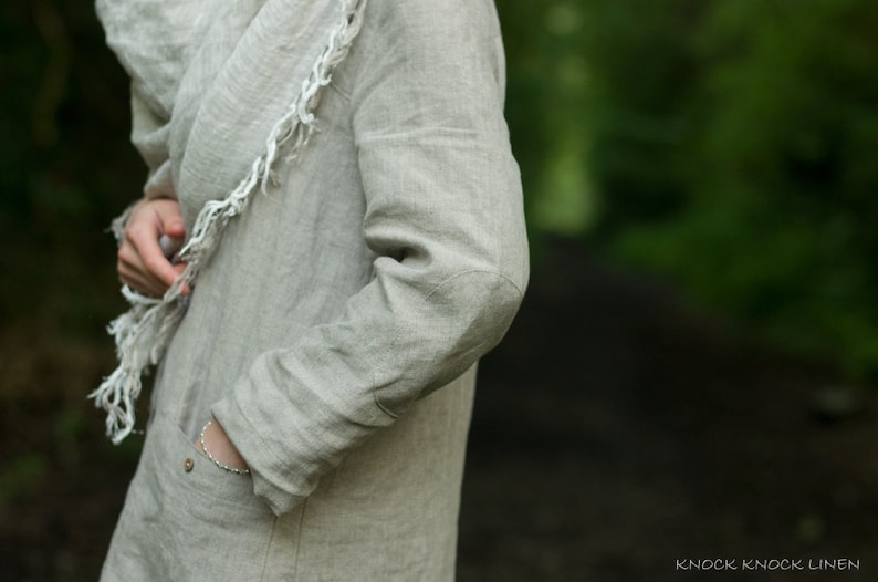 Close-up of a natural linen buttoned dress coat with long sleeves, worn by a person standing outdoors. One hand is tucked into a pocket; the outfit includes a matching linen scarf and a bracelet. Background features soft-focus greenery.
