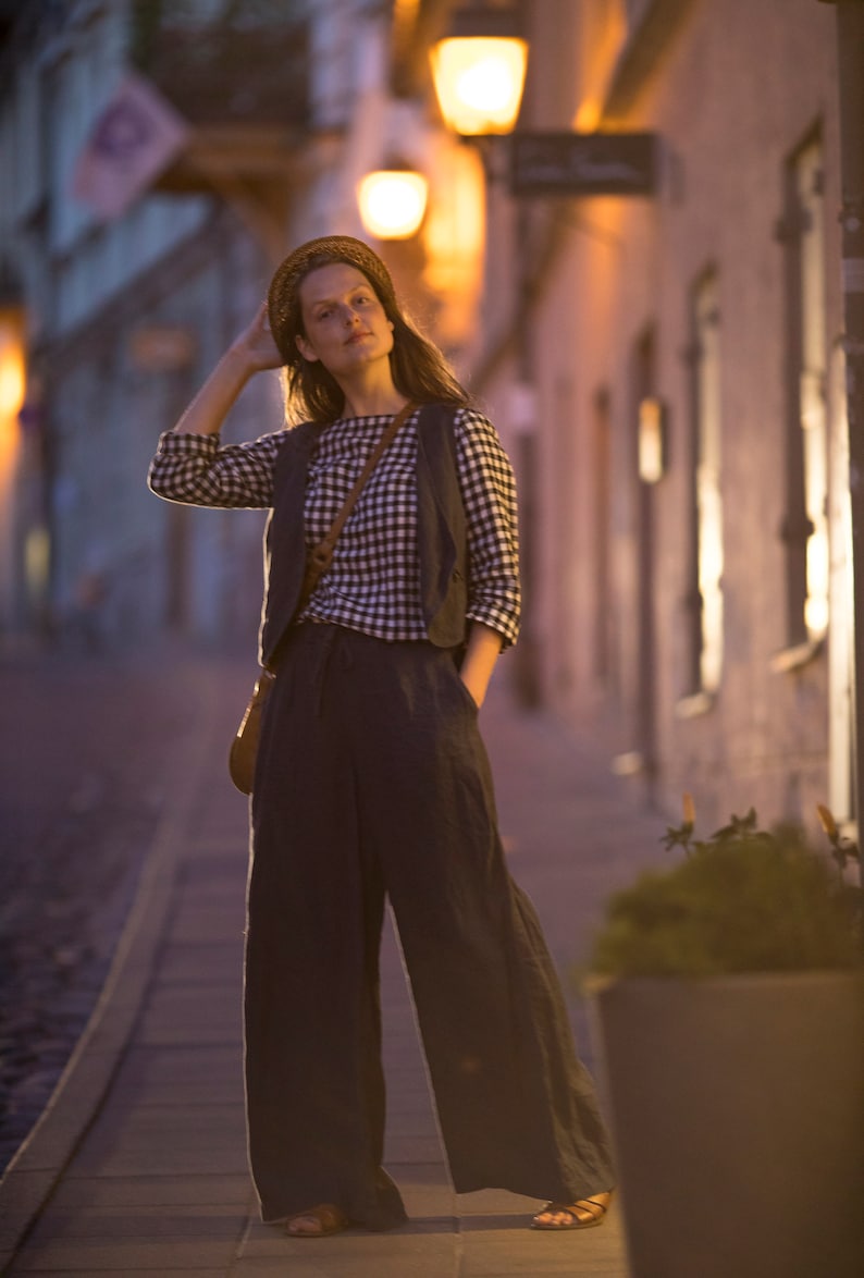 Charcoal linen wide-leg palazzo pants WIND on model, full-length front view; high waistline, loose drape with soft folds, one hand in side pocket, wide hems near sandals; dusk street lights blurred behind.
