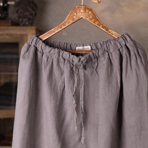 Gray linen skirt shown full-length on a hanger, front view, elasticated gathered waist with drawstring channel, dangling ties, wide A-line shape, plain front with no visible pockets, wooden cabinet and mannequins in background.