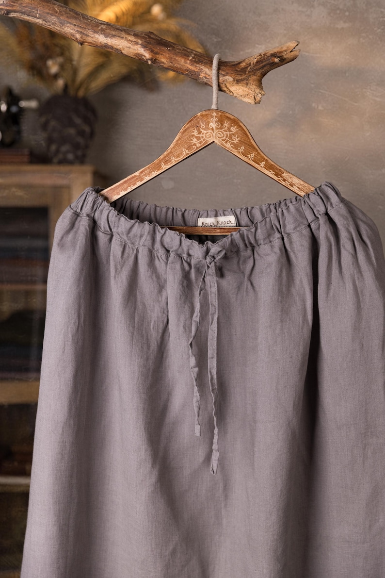 Gray linen skirt shown full-length on a hanger, front view, elasticated gathered waist with drawstring channel, dangling ties, wide A-line shape, plain front with no visible pockets, wooden cabinet and mannequins in background.