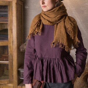 One loose dark purple linen blouse with long sleeves and a peplum ruffle at the waist, shown on a model standing indoors with a brown scarf wrapped around her neck and hands in grey trouser pockets.