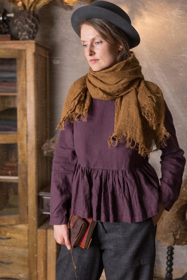 One loose dark purple linen blouse with long sleeves and a peplum ruffle at the waist, shown on a model standing indoors with a brown scarf wrapped around her neck and hands in grey trouser pockets.