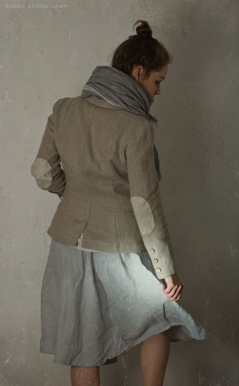 Grey-brown linen jacket worn by a model in a back three-quarter view, with curved back seams, pale elbow patches, long sleeves, cuff buttons, and a grey scarf around the neck.