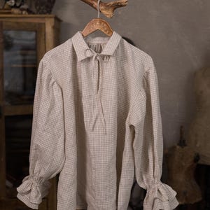 May include: A beige and white checkered long-sleeve shirt with a collared neckline and tie closure. The shirt has ruffled cuffs and is hanging on a wooden hanger. The shirt is made of a lightweight fabric.