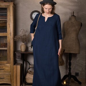 Front view of a loose navy long linen dress with 3/4 sleeves and simple kaftan neckline, shown on a model with hands in pockets in a vintage-style room.