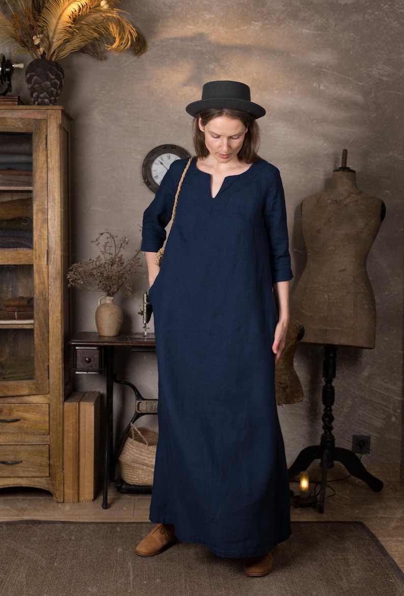 Front view of a loose navy long linen dress with 3/4 sleeves and simple kaftan neckline, shown on a model with hands in pockets in a vintage-style room.