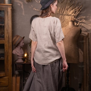 A young woman in a linen skirt and top stands in a room with vintage decor. She's wearing a hat and has her hands in her pockets. She's facing away from the camera.