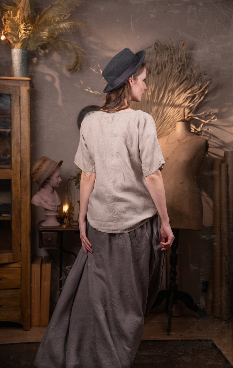 A young woman in a linen skirt and top stands in a room with vintage decor. She's wearing a hat and has her hands in her pockets. She's facing away from the camera.