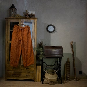Terracotta linen palazzo pants hanging on a hanger on a cabinet door, front view, with elastic waist, long drawstring ties, wide legs, visible pocket openings, creased linen texture, and a grey wall with a round clock behind.