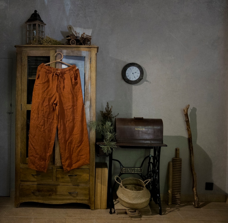 Terracotta linen palazzo pants hanging on a hanger on a cabinet door, front view, with elastic waist, long drawstring ties, wide legs, visible pocket openings, creased linen texture, and a grey wall with a round clock behind.