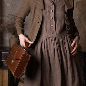 Close-up of woman wearing mushroom brown slim fit linen jacket unbuttoned over brown midi dress, showing curved collar, long sleeves and button front, holding brown leather bag in rustic studio, retro linen jacket blazer by knock knock linen.