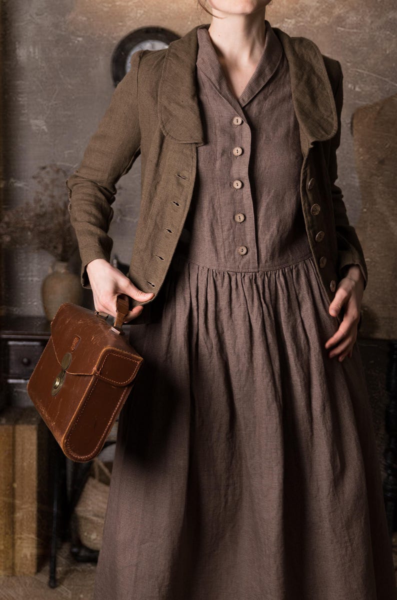 Close-up of woman wearing mushroom brown slim fit linen jacket unbuttoned over brown midi dress, showing curved collar, long sleeves and button front, holding brown leather bag in rustic studio, retro linen jacket blazer by knock knock linen.